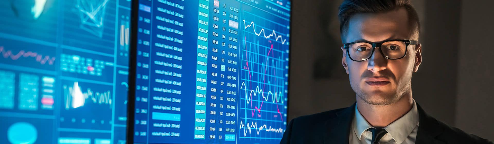 Un homme d’affaires portant des lunettes se tient devant deux grands écrans affichant des données et graphiques d’un logiciel maritime. Il regarde l’objectif en tenant une tablette numérique.