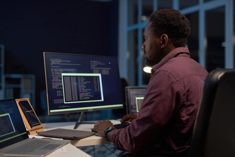 ein Mitarbeiter afrikanischer Abstammung sitzt an einer Computertastatur an einem Schreibtisch. Der Computermonitor befindet sich neben einem Smartphone, einem Tablet mit Case und einem Laptop.