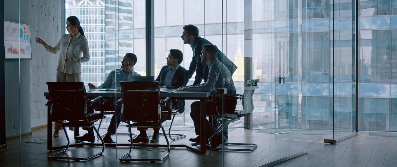 a glass-walled office with a female professional pointing to maritime shipping data and graphs on a wall monitor as three businessmen sit a table looking on. A fourth is standing also looking on.