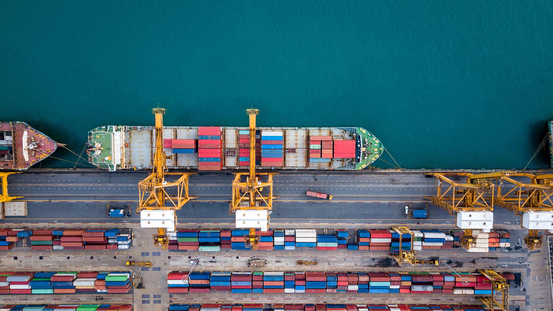 cargo container ships at port seen from aloft. One ship is the center of unloading activity with cranes overhanging it and a yard of shipping containers behind them.