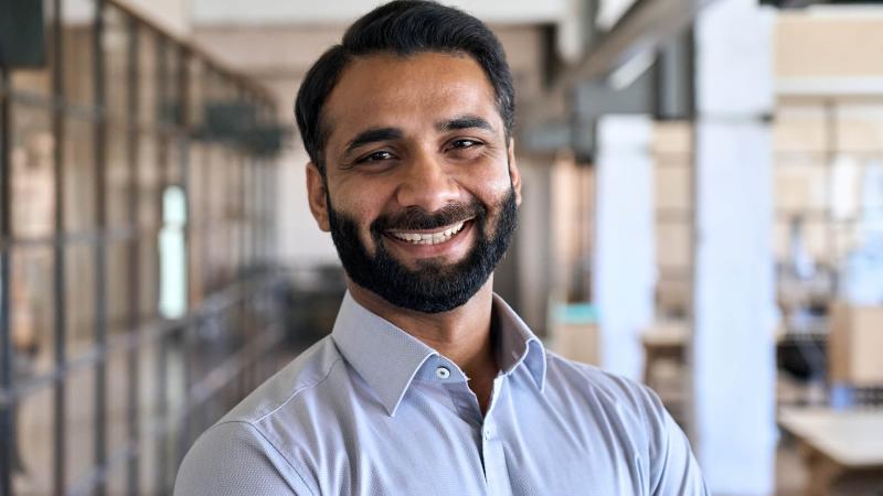 a bearded man of Indian descent stands smiling looking at the camera as he stands in a common area of a maritime shipping office. 