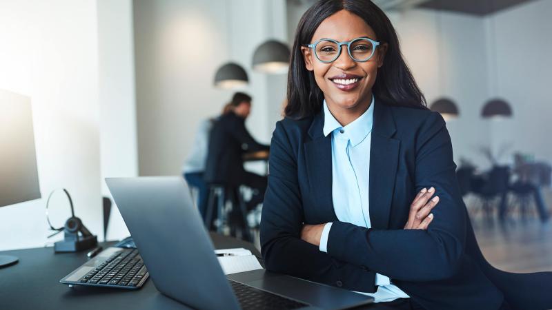 a businesswoman of African descent sits at a desk smiling looking at the camera. She's wearing glasses and smiling as she sits at her desk in a maritime shipping office. 