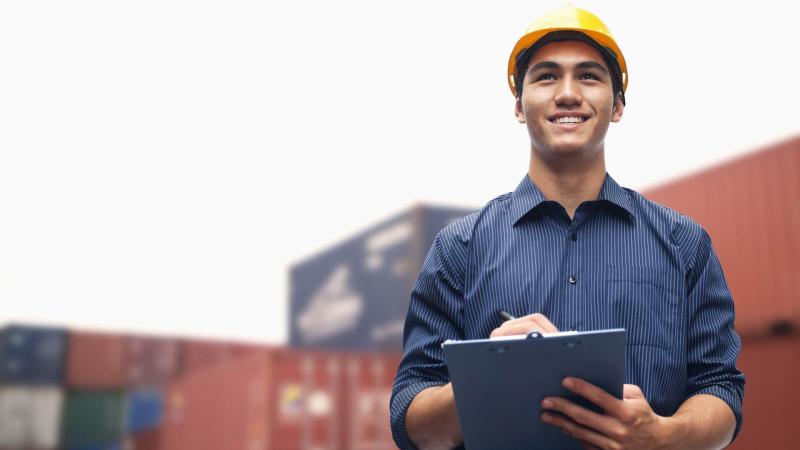 a young smiling man wearing a hard hat writing on a clipboard  amidst shipping containers in  a maritime shipping terminal. 