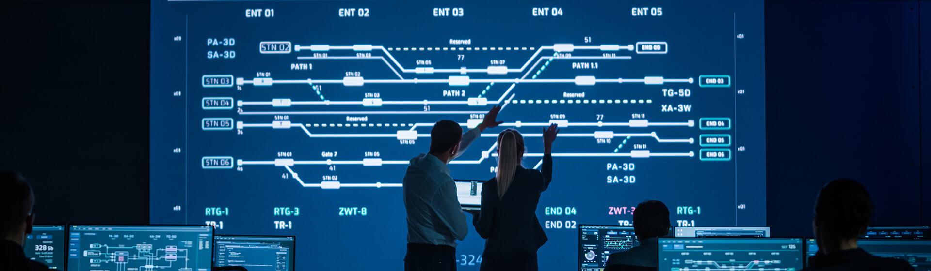 a professional man and woman in a control room interacting with a large monitoring screen showing rail routes at a maritime shipping terminal.