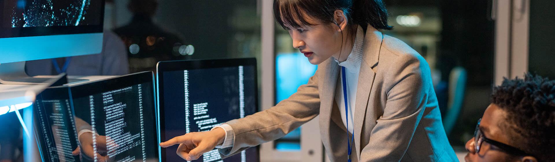 a young woman of asian descent IT engineer points at a one of three computer monitors while a young man of african descent sits at his desk at the keboard. 