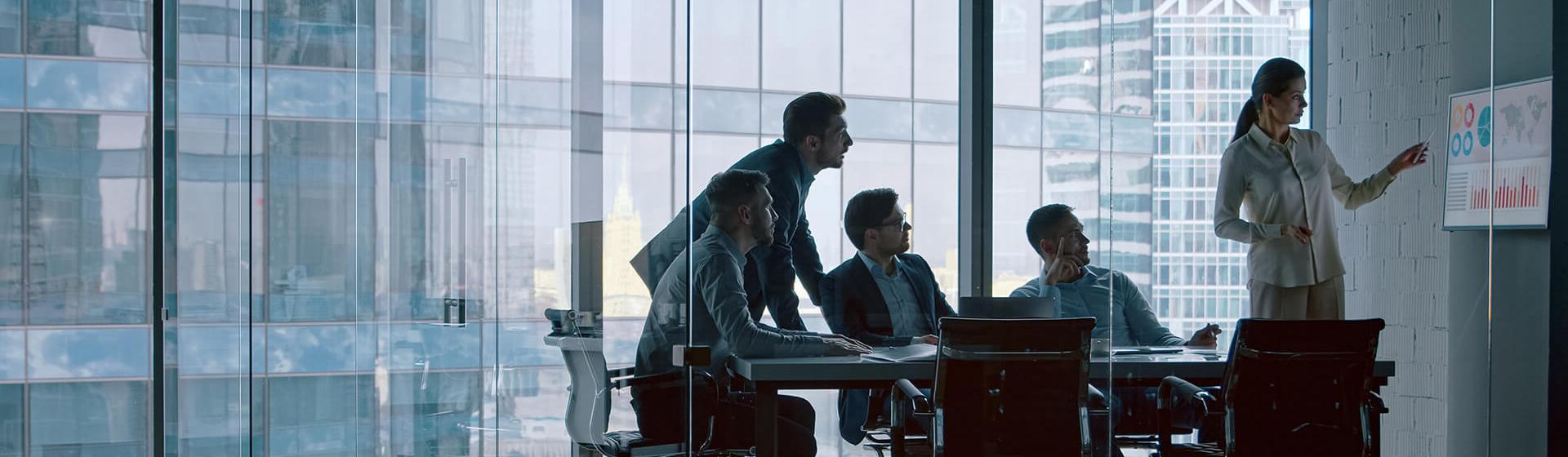 Een kantoor met glazen muren met een vrouwelijke professional die zeevaartgegevens en -grafieken op een monitor aanwijst en drie zakenlieden die aan tafel toekijken. Een vierde persoon staat en kijkt ook.