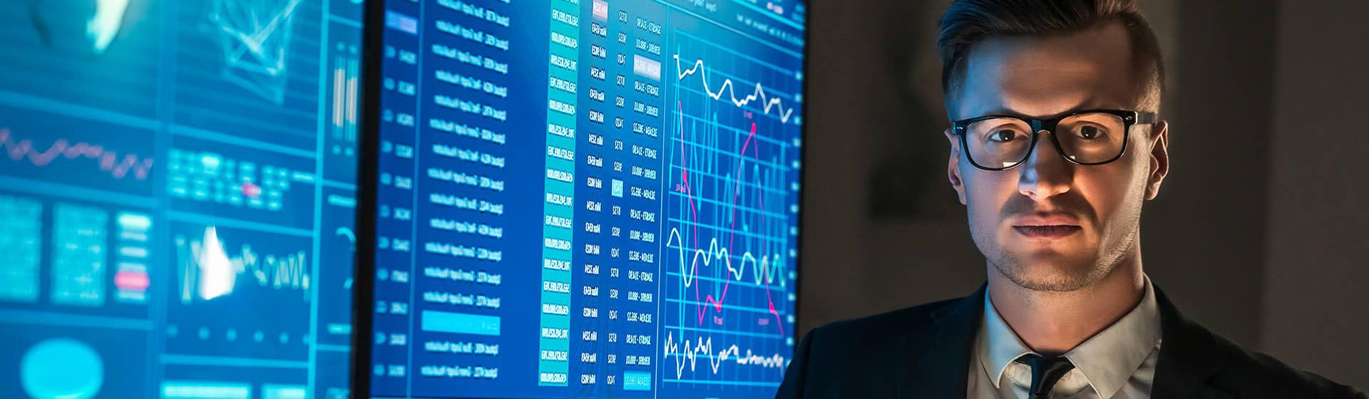 a businessman wearing glasses stands in front of tow large monitors showing maritime software data and graphs. He looks into the camera holding a digital tablet. 