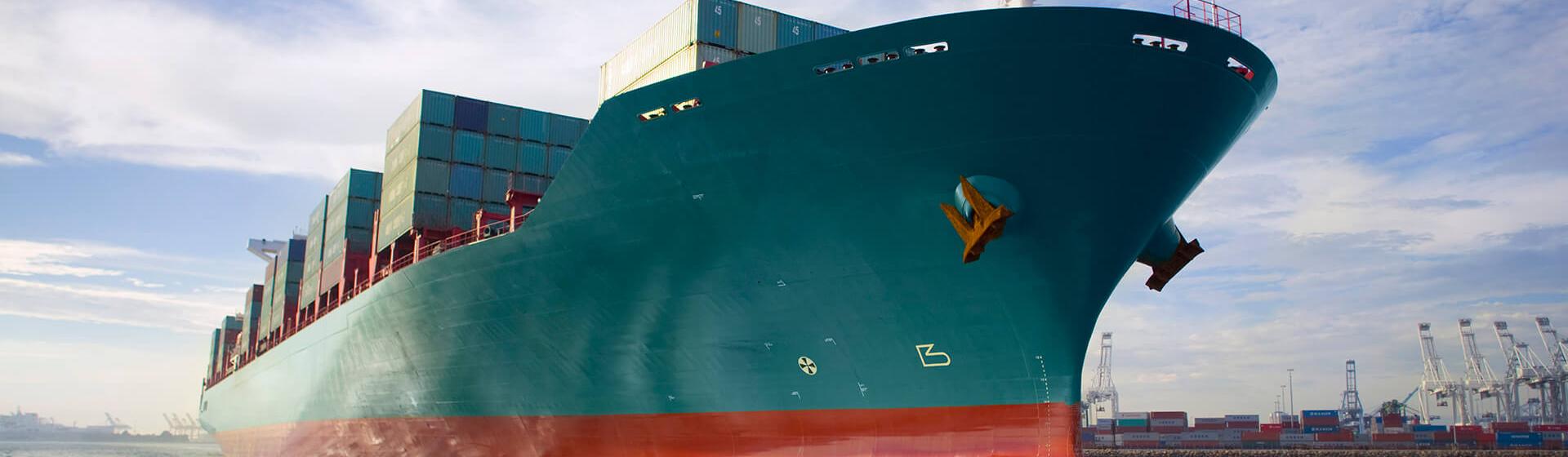 a cargo container ship moving through a harbor port with cranes in background as seen from sea level.