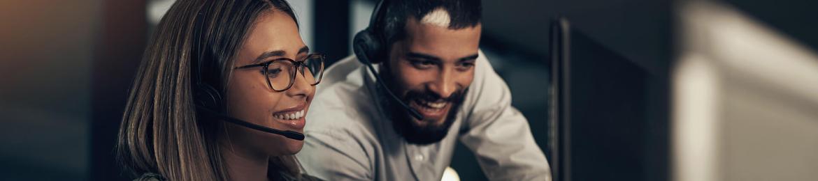 zwei Büroangestellte mit Kopfhörern, die lächelnd um einen Schreibtisch versammelt die verbesserten Ergebnisse der Schifffahrtsbranche auf einem Computerbildschirm prüfen.