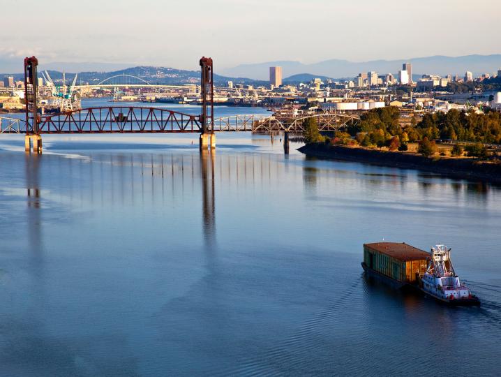 a tugboat pushing a river barge seen from aloft two points off port stern along a river with multiple bridges amidst an urban and park setting.