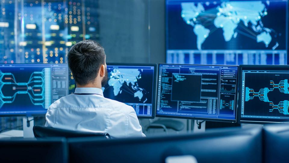 a man sits at a desk, surrounded by computer monitors, with his back to the camera. He's looking to a large screen displaying a map on a far wall. 