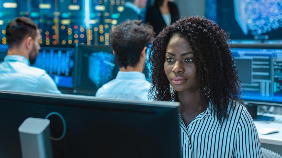 一位非洲裔女士坐在电脑显示器前。背景中的其他专业人员坐在各自的电脑前。
