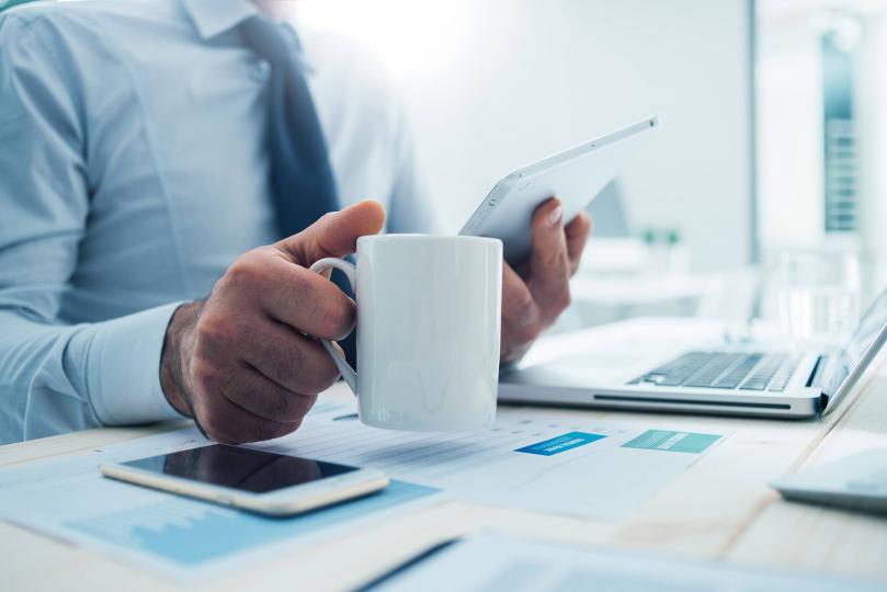 Un homme, une tasse de café à la main, regarde une tablette.