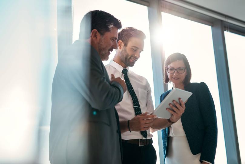 Three maritime professionals, standing in front of floor to ceiling windows, looking at single digital tablet smiling at purchasing results. 